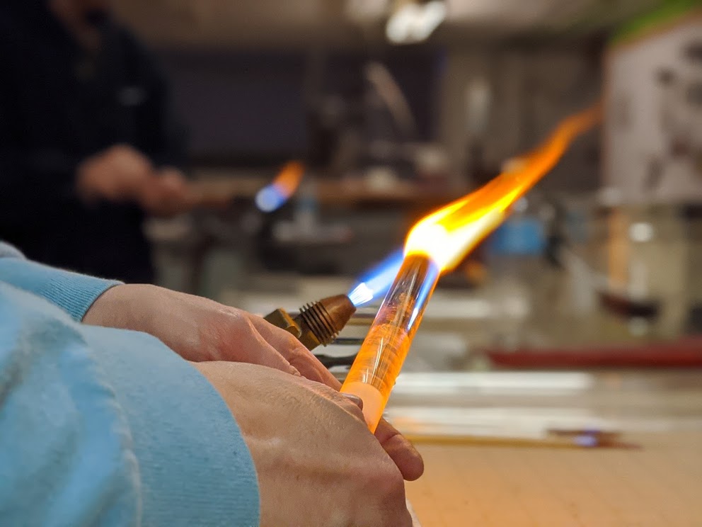 A student heats up a large piece of glass in-flame, the reflection seemingly make the glass glow orange.