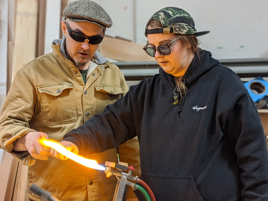 John guides a student's hand in the flame to help her effectively roll the glass so pulling a rod becomes much easier.