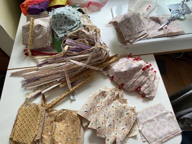 An overhead view of the process of sewing hundreds of masks by hand, including the pleat markers, half-sewn cloth masks, and excess materials on Anchorage Makerspace member's desk.